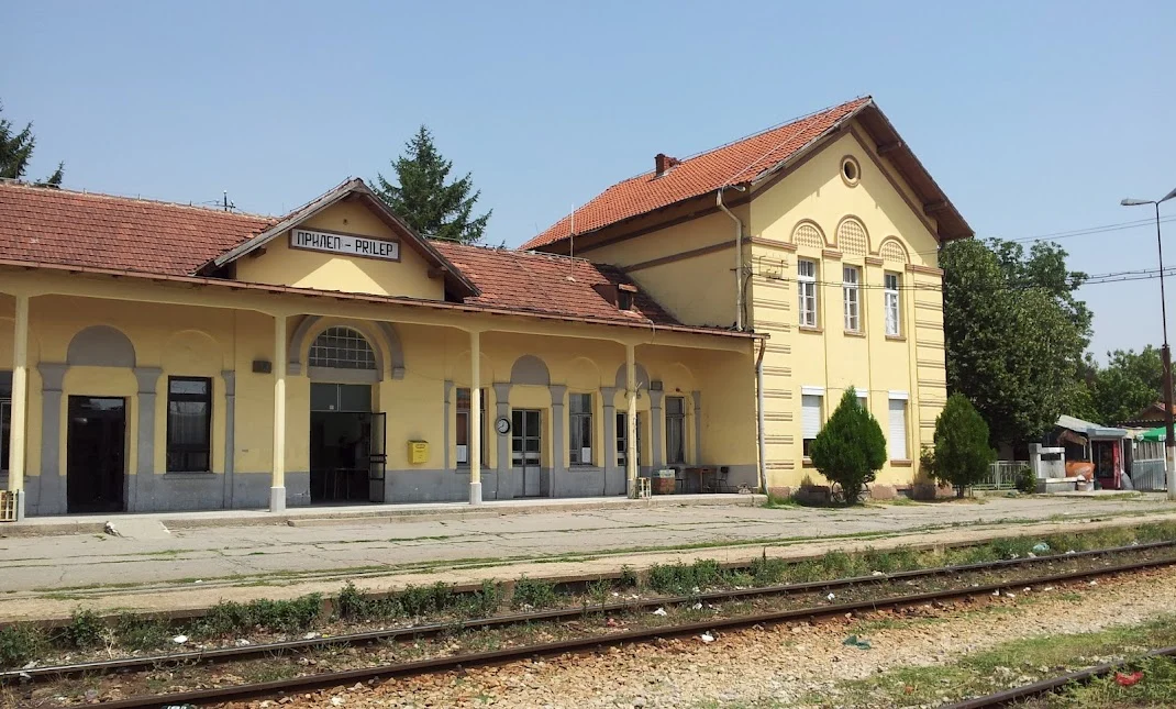 The yellow station building of Prilep, with a red-tiled roof and a Prilep sign above the entrance, seen across the platform and tracks on a sunny day