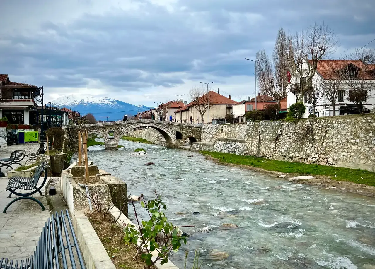 The Lumbardhi river running through central Prizren between stone embankments, with the Stone Bridge and snowy mountains in the distance