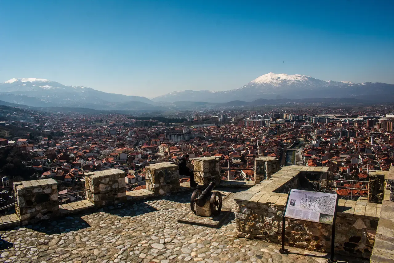The stone terrace of Prizren Fortress with old cannon and an information board, and the town and snow-capped mountains below