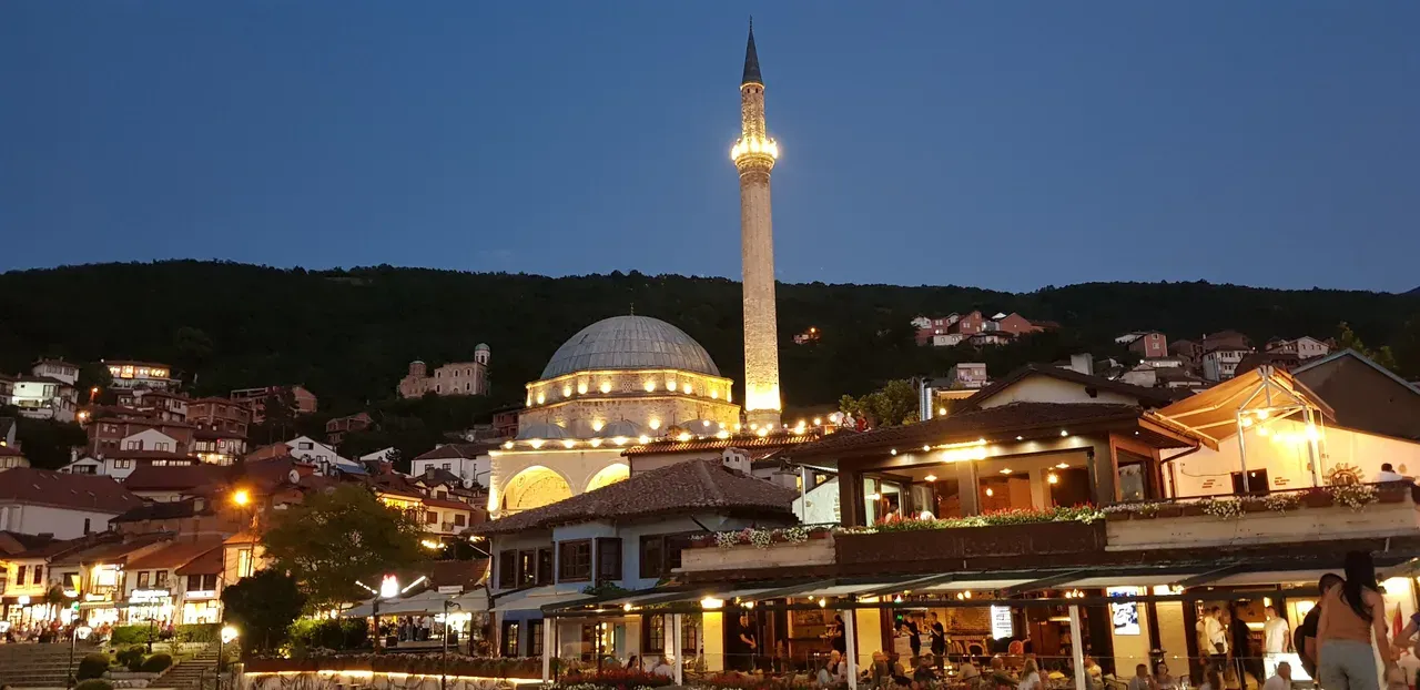 Sinan Pasha Mosque and its minaret lit at dusk, with the lights of riverside cafes in old-town Prizren