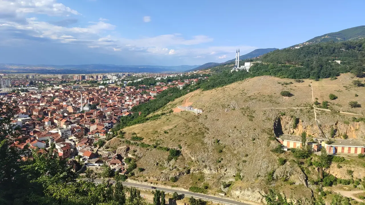 Red roofs of Prizren spreading across the valley below a bare hillside, with a minaret and mountains on the horizon
