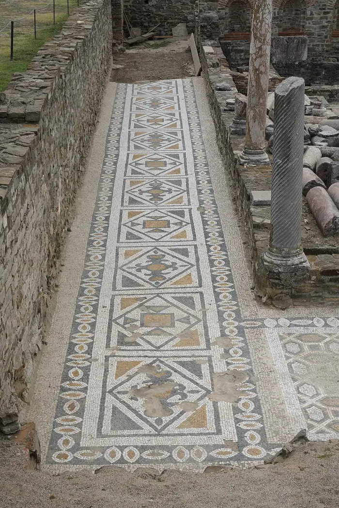 A long geometric mosaic pavement running between walls at Stobi, with a fallen column beside it