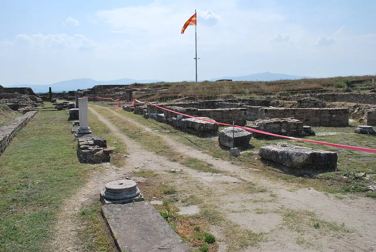 A paved ancient street at Stobi lined with column bases and low walls, with a flagpole beyond