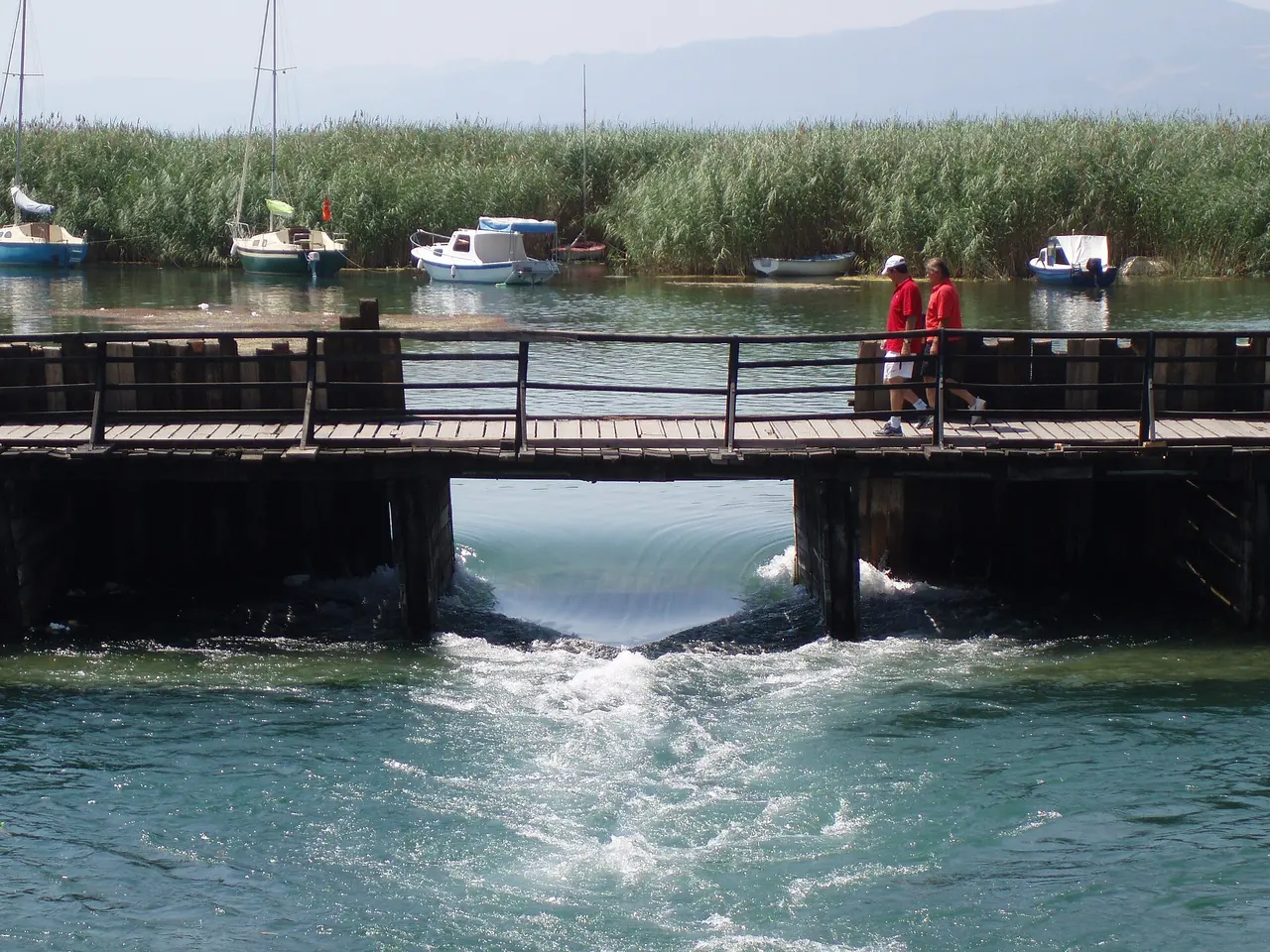 Pedestrian Bridge of the Poets over the clear Black Drin at Lake Ohrid with hills beyond
