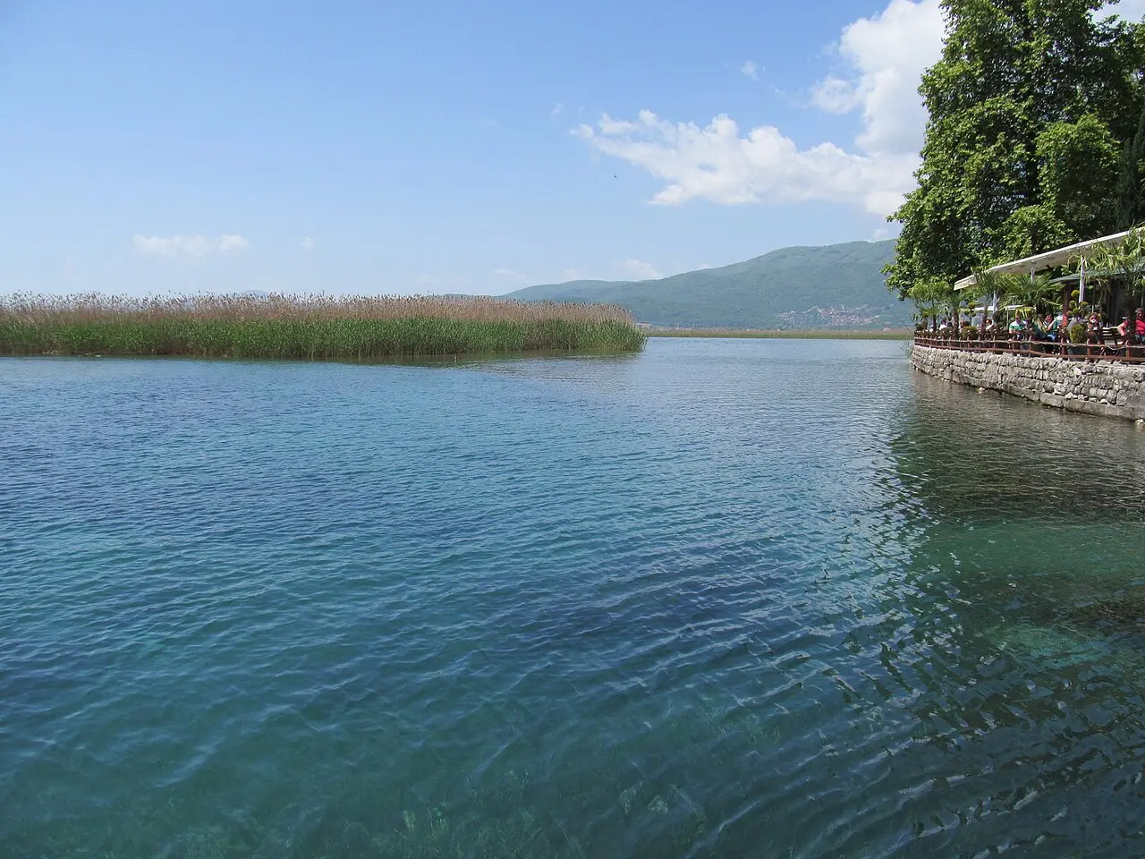 Clear blue-green water of Lake Ohrid beside a stone-walled café terrace in Struga, with reed beds and hills across the bay