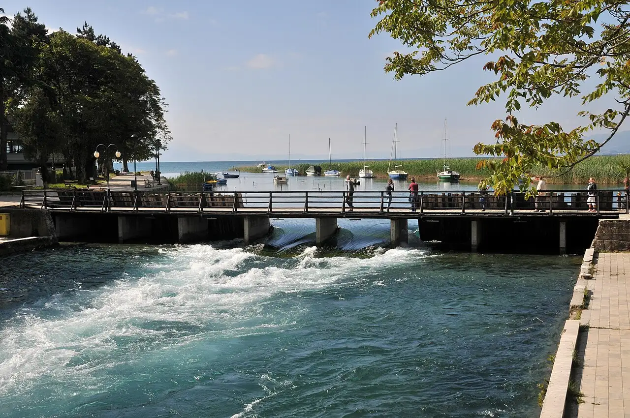 People standing on the wooden needle dam over the Black Drin outflow in Struga, with boats and Lake Ohrid behind