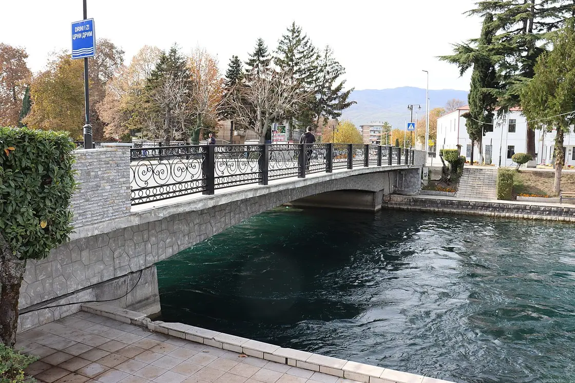 An ornate pedestrian bridge with decorative iron railings over the emerald Black Drin, signed Drini i Zi / Crn Drim, in Struga