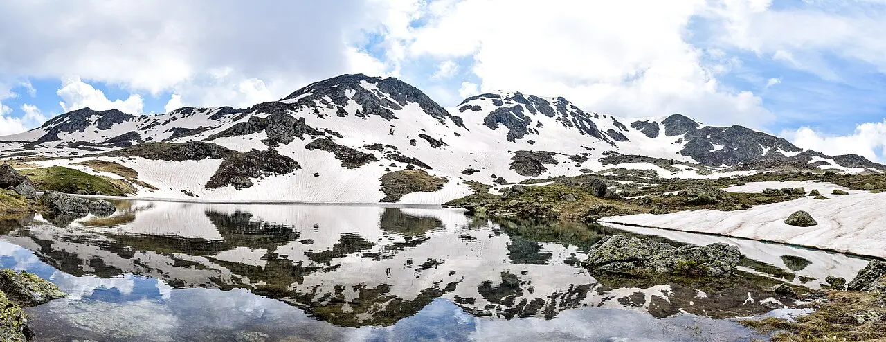 A still mountain lake reflecting snow-streaked peaks under a cloudy sky in the hills above Vevčani near Struga