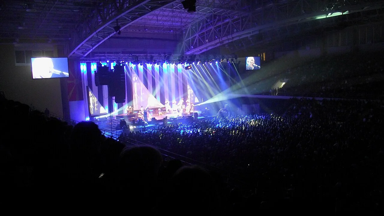 A packed concert at Boris Trajkovski Arena in Skopje, the crowd lit blue in front of a stage with light beams and two video screens