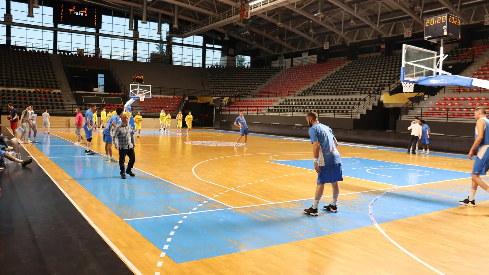 Basketball players warming up on the wooden court of the Jane Sandanski Arena in Skopje, below red and grey tiered seating and a steel roof
