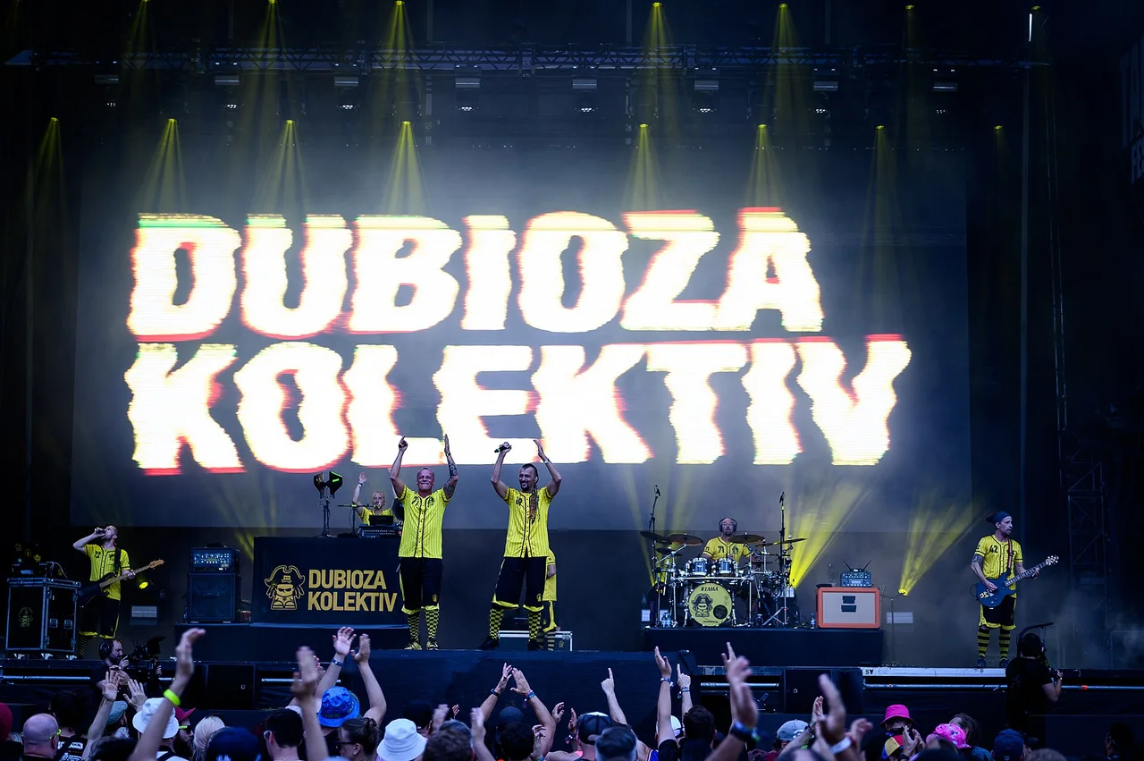 Dubioza Kolektiv on an outdoor festival stage in yellow shirts, arms raised under a huge screen showing the band's name, with the crowd's hands in the foreground
