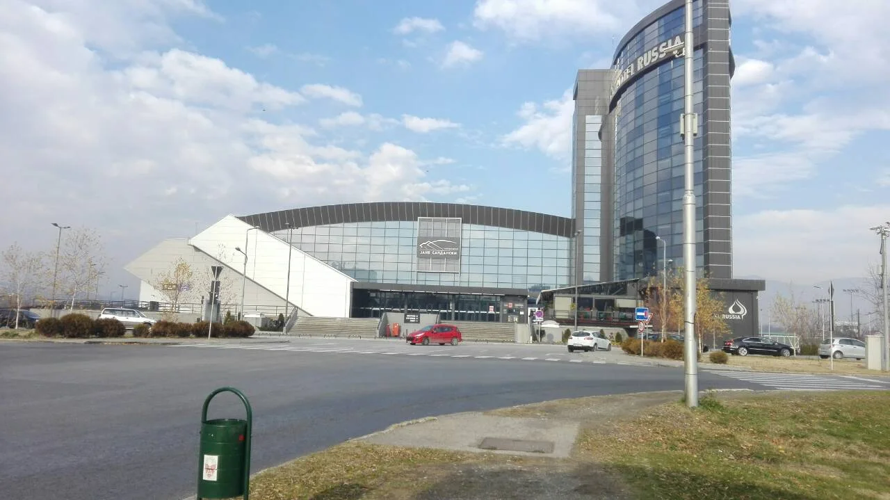 The curved glass front of the Jane Sandanski sports centre in Skopje with the tall glass Hotel Russia tower beside it and a wide road in front