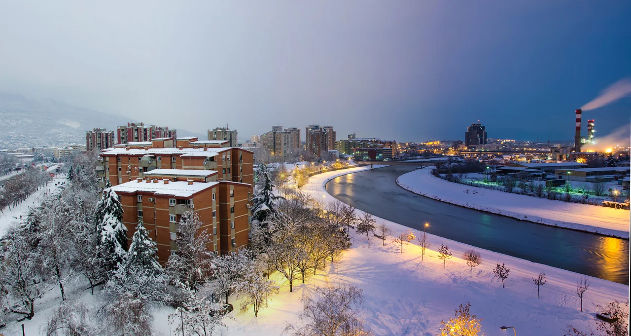 Skopje under snow at dusk, with apartment blocks, snow-covered trees and lit riverside paths along the curving Vardar river