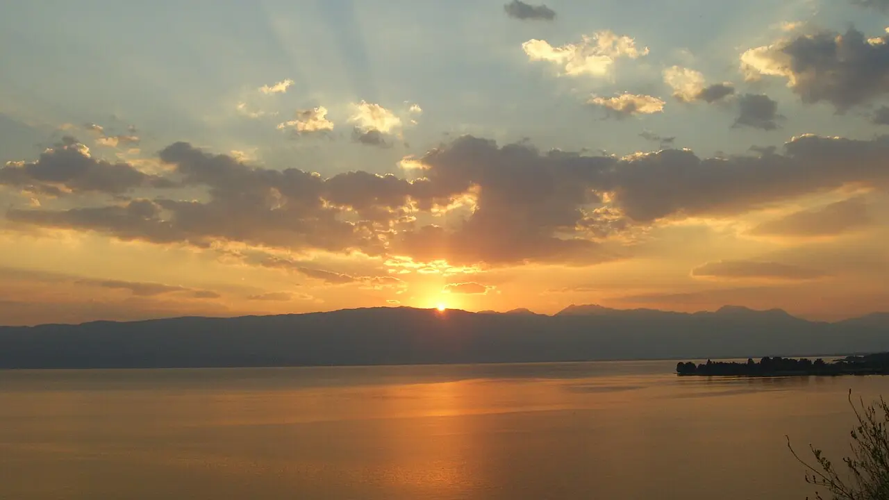 A dramatic sunset over Lake Ohrid, with sun rays breaking through clouds above the water