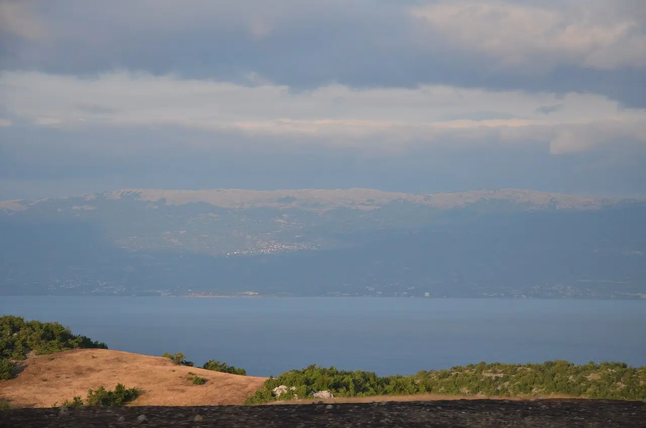 A wide view over the still water of Lake Ohrid toward hazy hills and a distant lakeside town