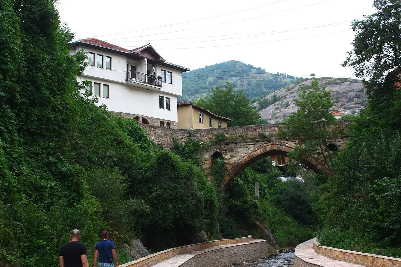 A single-arch Ottoman stone bridge over the wooded gorge in Kratovo, with a white house above and two people walking below
