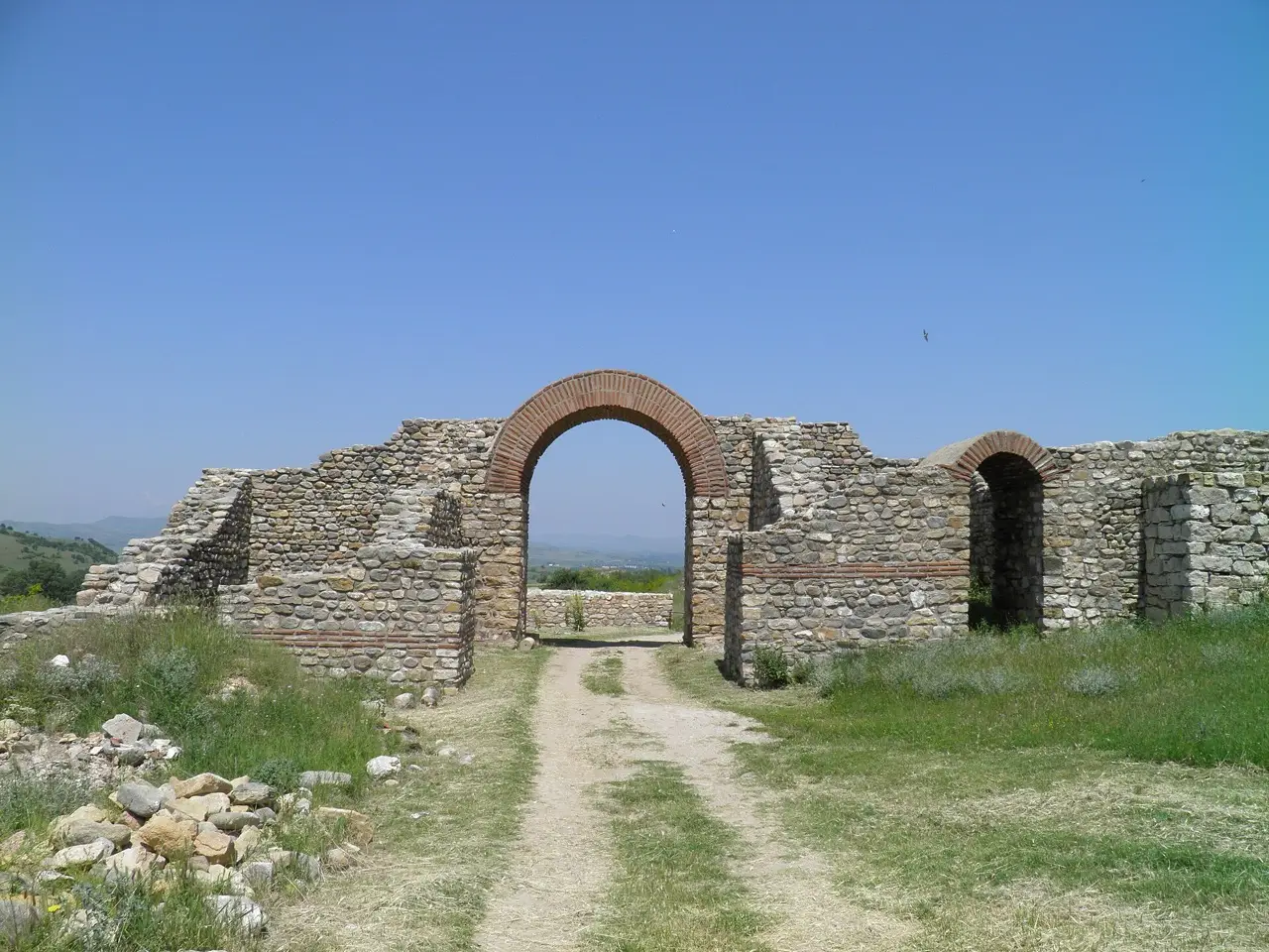 The restored arched main gate of the Bargala archaeological site, framed against a clear blue sky