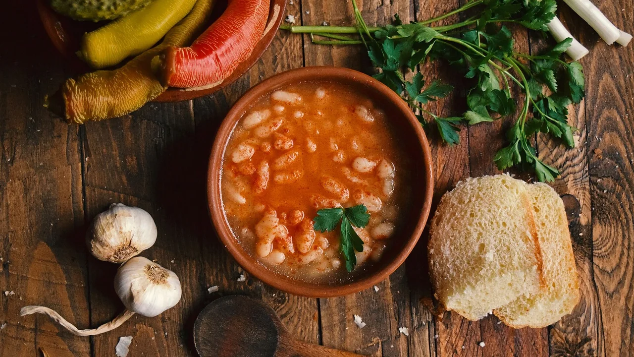 A clay bowl of white bean soup with paprika beside pickled peppers, garlic, parsley and bread on a wooden table
