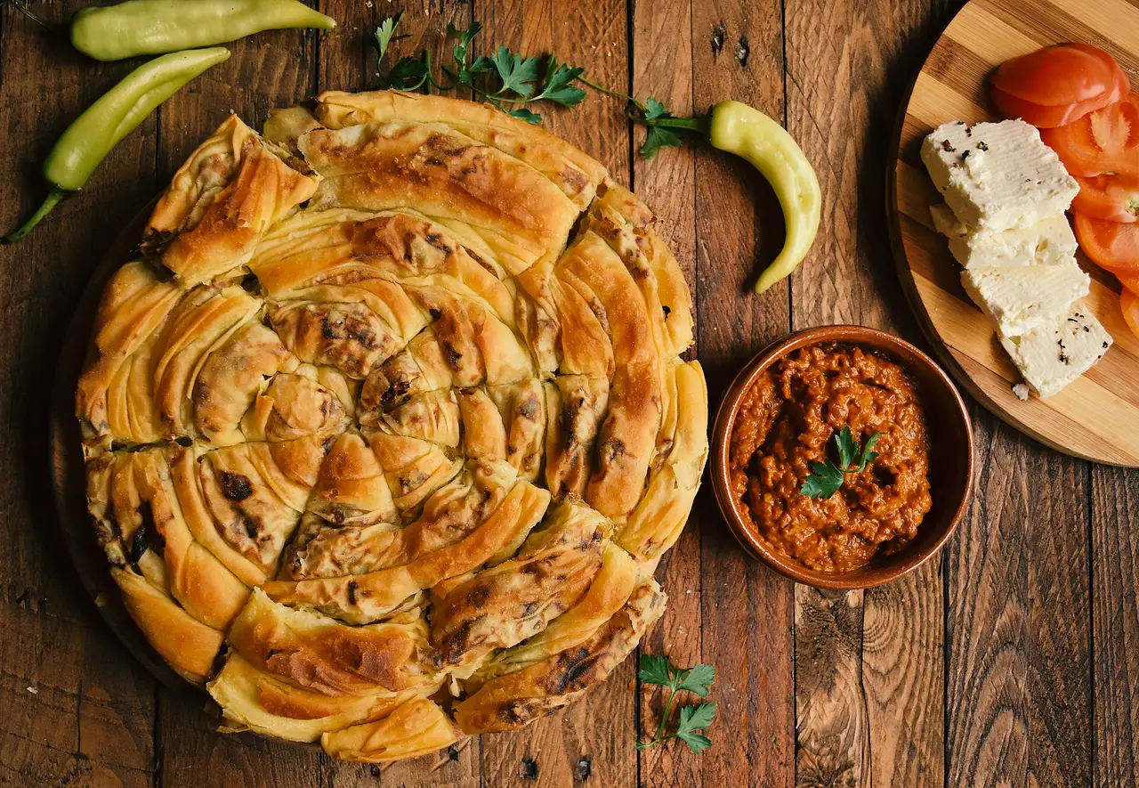 A round spiral leek zelnik pie on a wooden board beside a bowl of red pepper spread, slices of white cheese and tomato, and green peppers