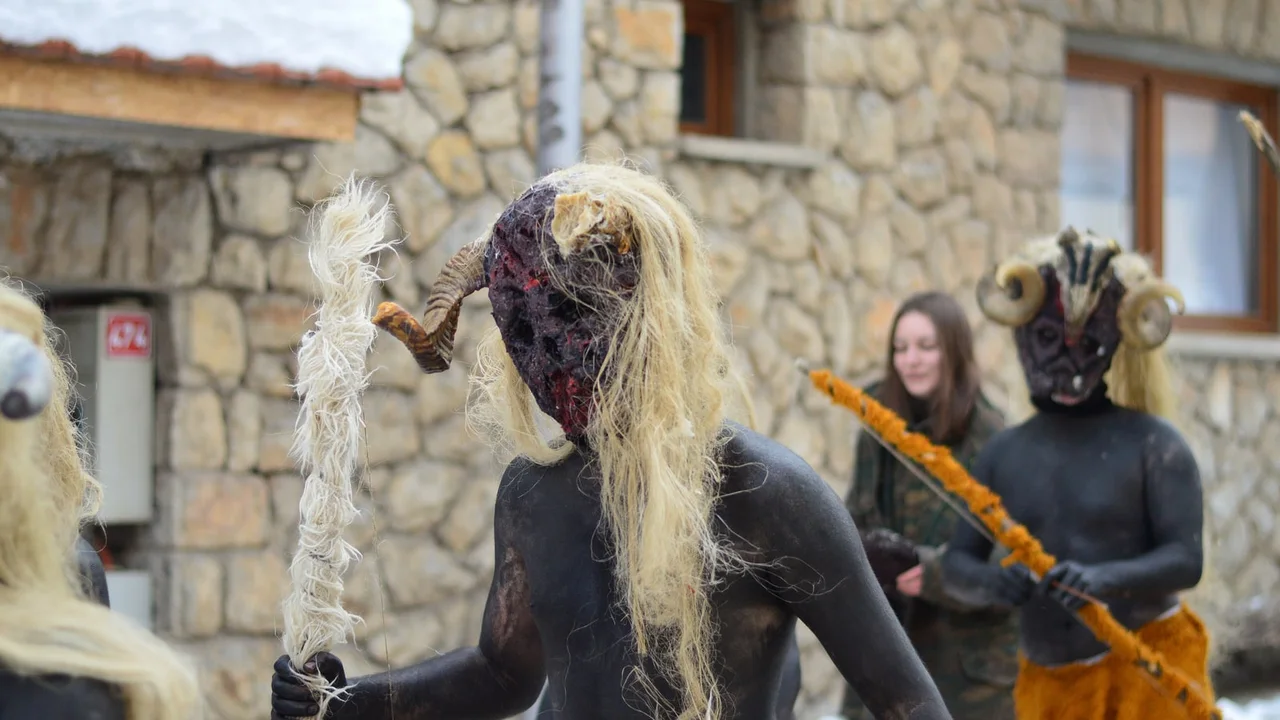 A Vevčani carnival mask in winter: a figure with a dark painted body, long pale hair and curled horns, with another masked figure behind
