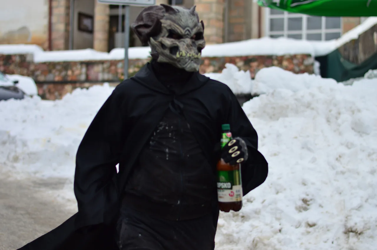 A figure in a black cloak and a grey monster mask carrying a bottle in the snow in Vevčani