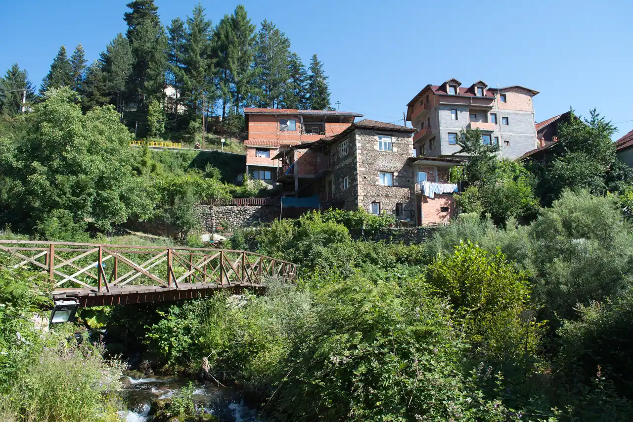 Stone and brick houses of Vevčani on a wooded slope above a stream and a wooden footbridge