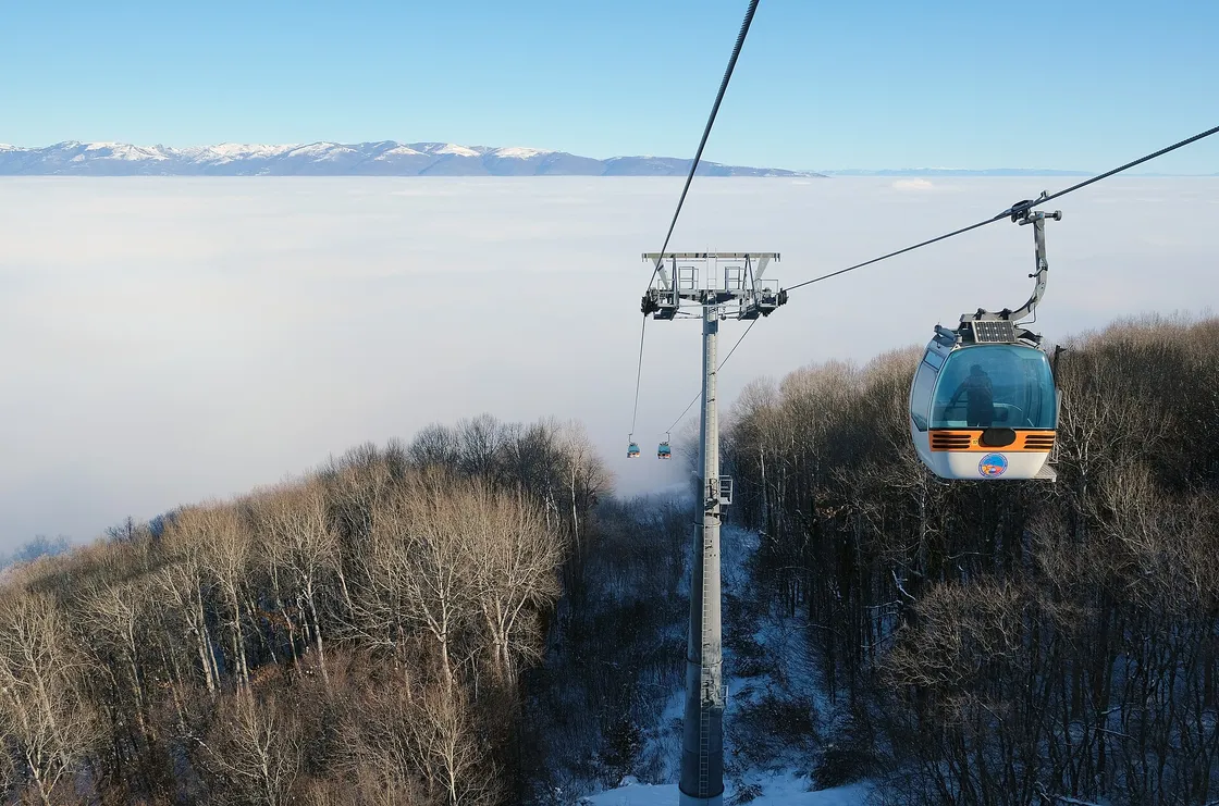 A cable car gondola climbing above Vodno's forest with a low cloud sea over Skopje and distant mountains