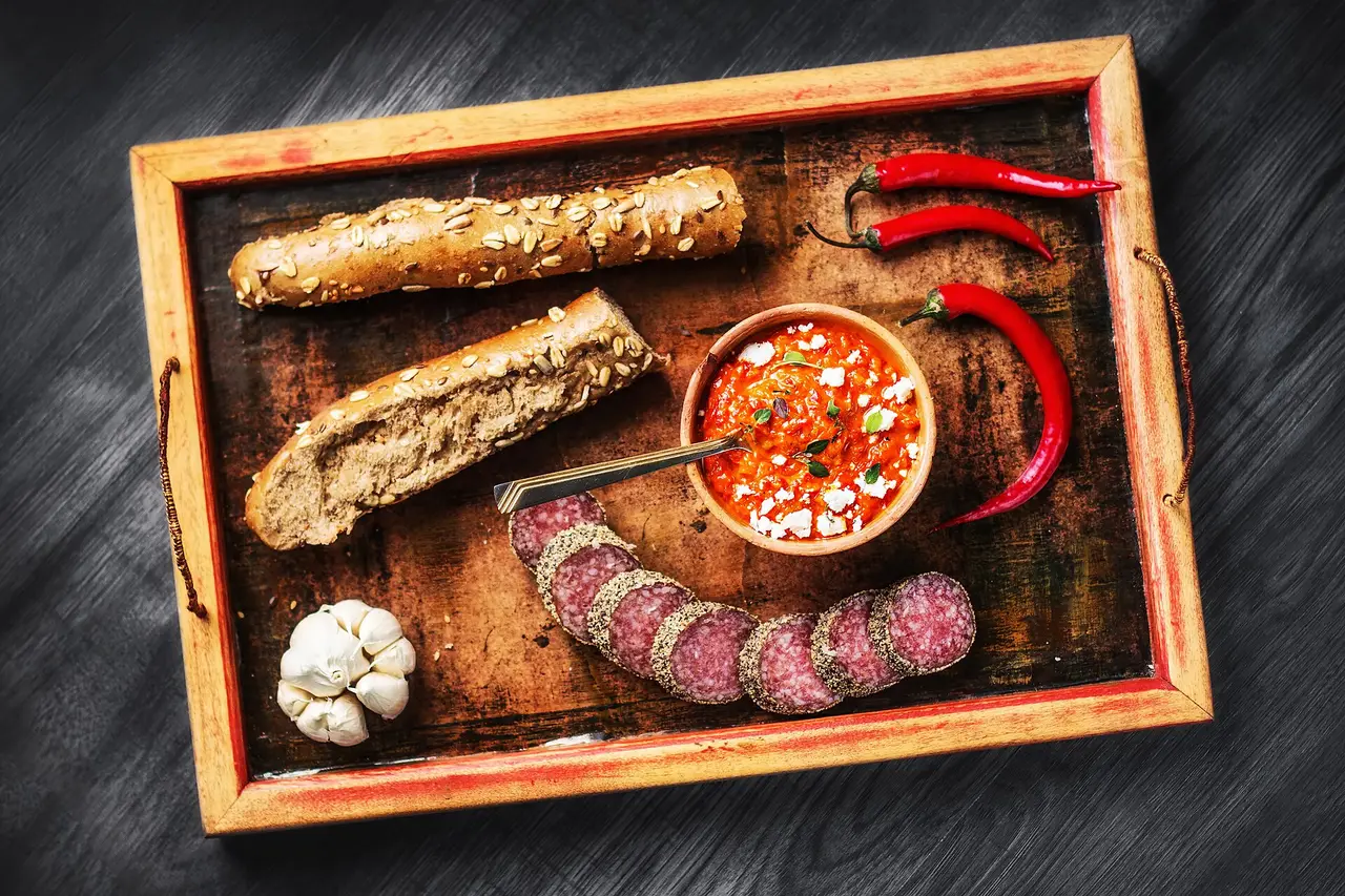 A bowl of ajvar topped with crumbled white cheese, served with bread, cured sausage and chilli peppers