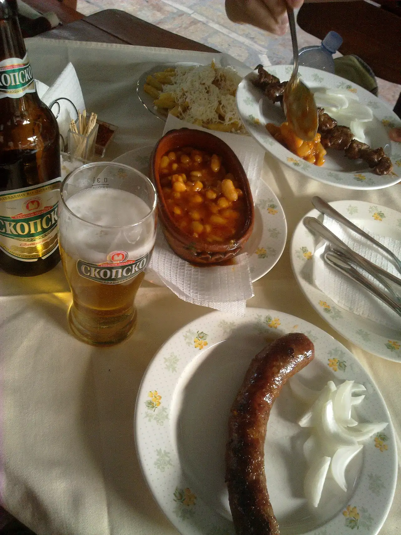 A kafana table with a bottle of Skopsko beer, tavče gravče in a clay pot, grilled meat and a sausage