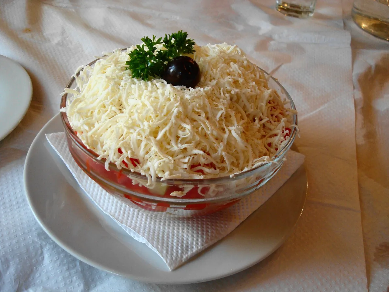 A bowl of šopska salad topped with a thick mound of grated white cheese, parsley and an olive