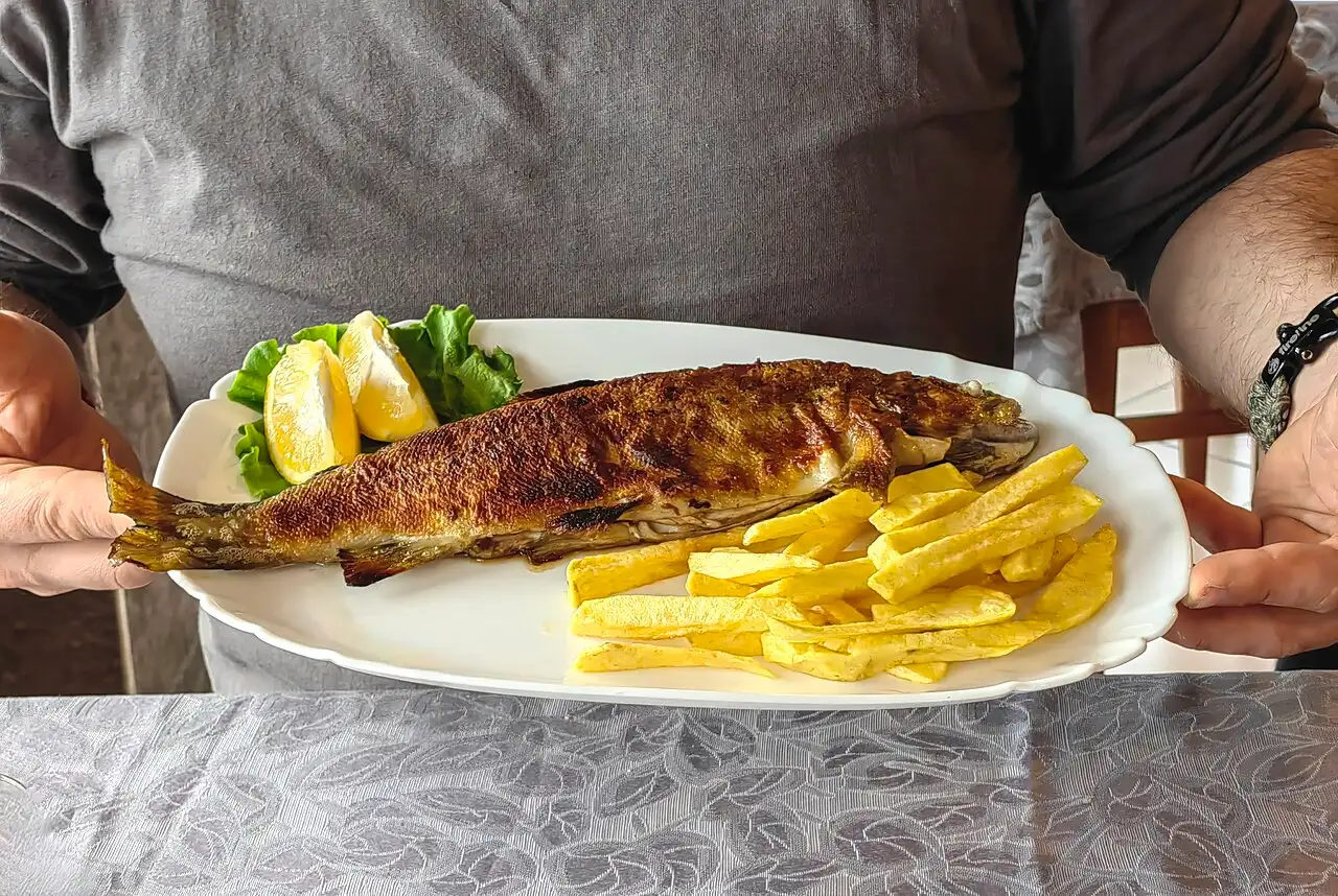 A whole grilled Ohrid trout served on a white platter with chips, lettuce and lemon wedges