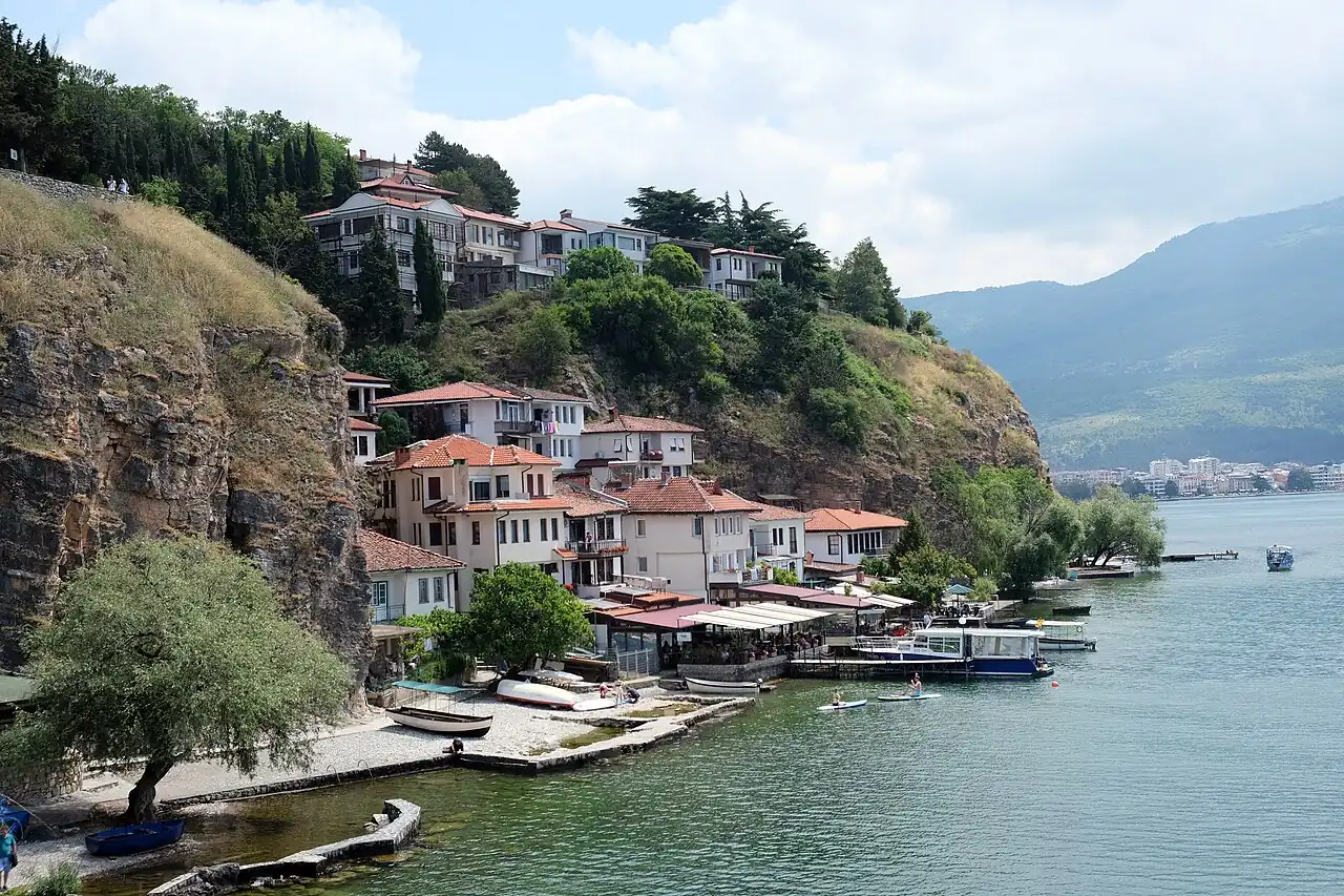 Fish restaurant terraces with white awnings lining the Kaneo cove on Lake Ohrid, boats moored and swimmers in the clear water below the cliff houses