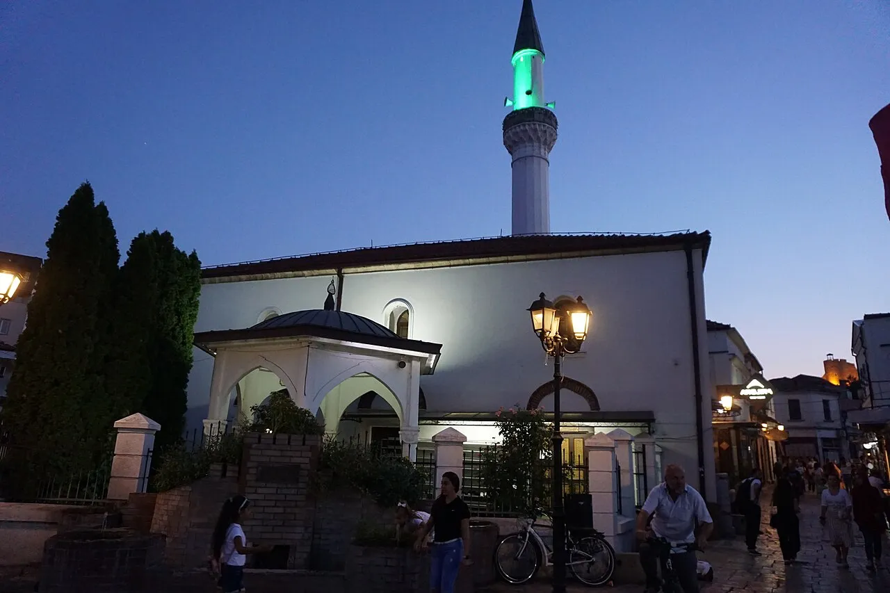 A white mosque with a green-lit minaret in Skopje Old Bazaar at dusk, with people walking