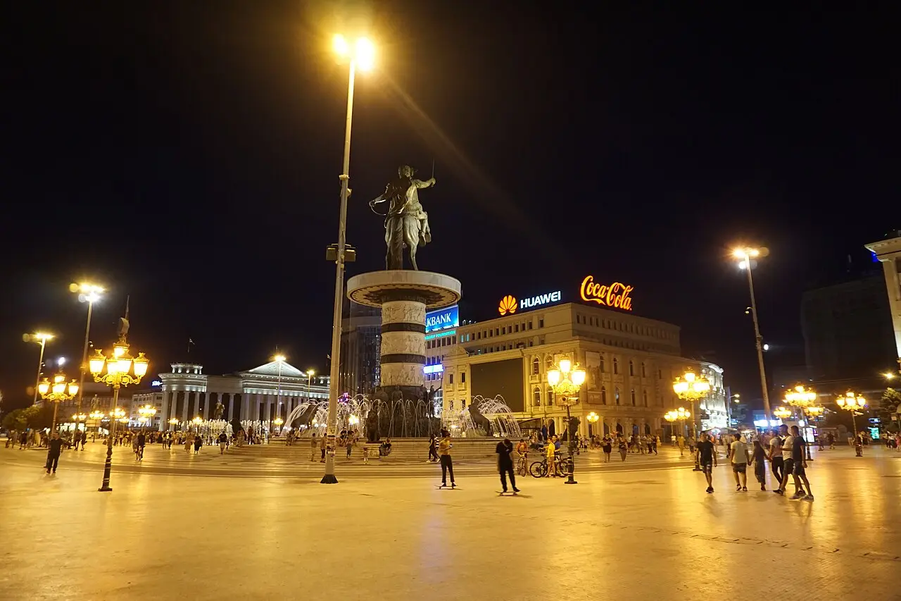 Wide view of Macedonia Square in Skopje at night with the floodlit Warrior on a Horse fountain and people crossing the square