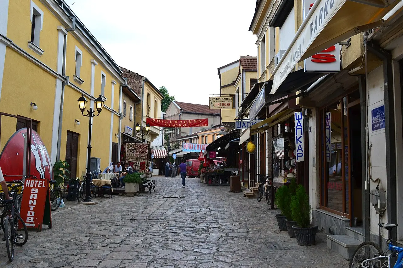 A cobbled lane in the Old Bazaar of Skopje lined with shops and cafés, with a small hotel sign and the hillside beyond