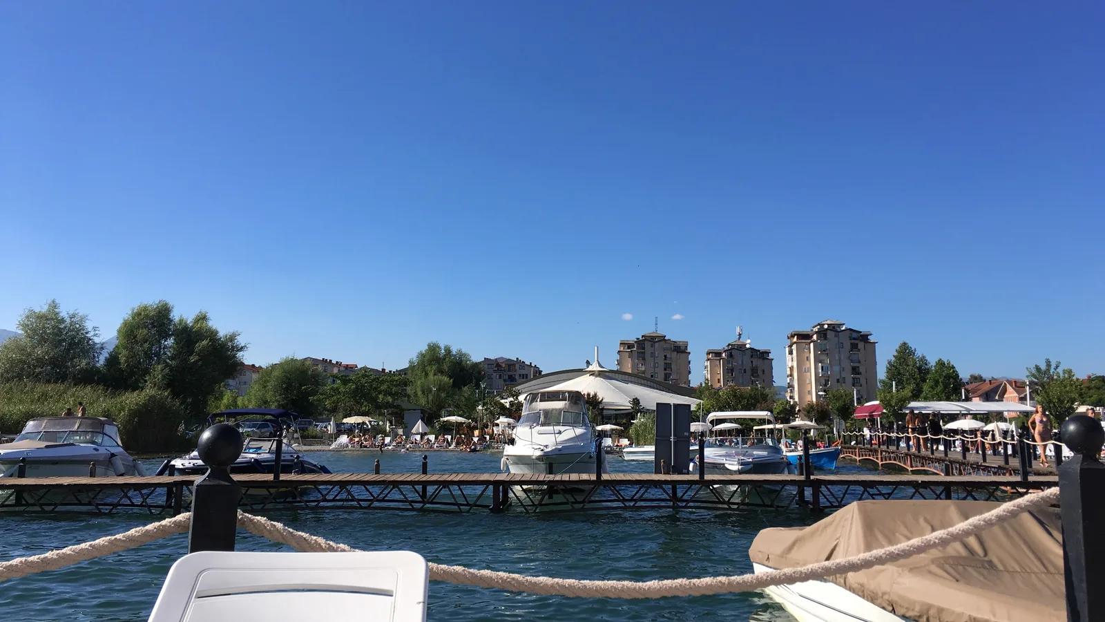 Wooden jetty with motorboats in front of a beach bar and apartment blocks on the Struga lakefront in summer